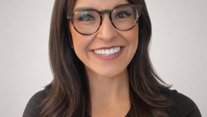 A woman with long dark hair and glasses smiles warmly against a plain light gray background.