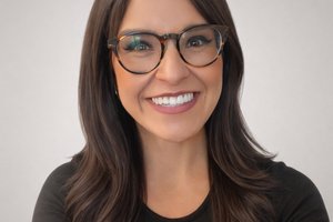 A woman with long dark hair and glasses smiles warmly against a plain light gray background.
