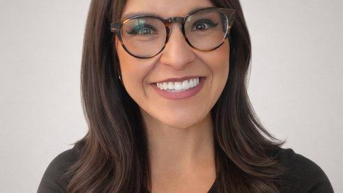 A woman with long dark hair and glasses smiles warmly against a plain light gray background.