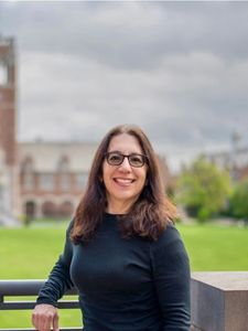 A woman with long brown hair and glasses smiles while leaning on a railing with a large brick building in the background.