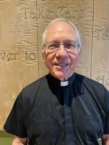 An older man wearing glasses and a black clerical shirt with a white collar stands smiling in front of a carved wooden wall.