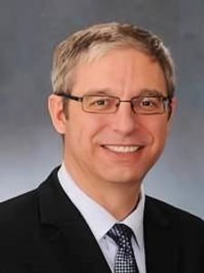 A middle-aged man with short gray hair and glasses smiles while wearing a dark suit, white shirt, and patterned tie.