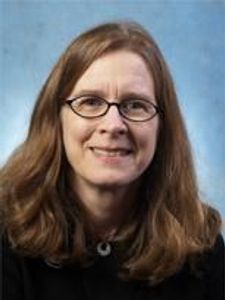 A woman with shoulder-length brown hair and glasses smiles gently against a plain blue-gray background.