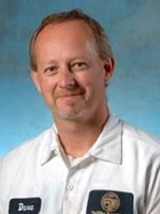 A man with short light brown hair and a goatee smiles slightly while wearing a white lab coat with a name tag that says Dave.