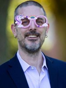 A man with short dark hair and glasses wearing a light blue button-up shirt stands against a blurred green background.