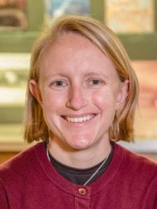 A person with short blonde hair smiles directly at the camera wearing a maroon sweater and black shirt.