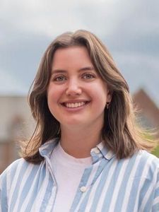 A young woman with shoulder-length brown hair smiles at the camera against a light gray background.