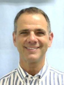 A middle-aged man with short dark hair smiles at the camera wearing a white and blue striped collared shirt.