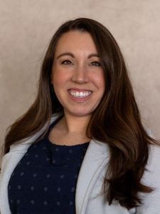 A woman with long brown hair smiles while wearing a dark blue top and light gray blazer against a plain background.