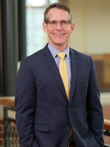 A man wearing glasses, a dark suit, striped shirt, and yellow tie is smiling indoors near a railing.