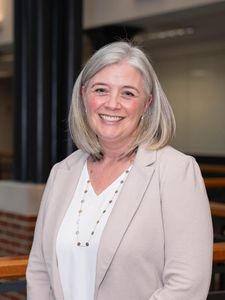 A smiling woman with shoulder-length gray hair wearing a light beige blazer and white top stands indoors.
