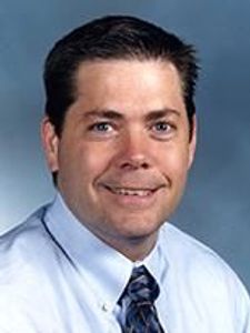 A man with short dark hair wearing a white shirt and patterned tie smiles against a plain blue-gray background.
