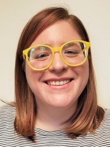 A person with shoulder-length light brown hair smiles at the camera wearing large yellow glasses and a striped shirt.