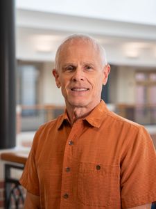 A smiling older man with short white hair wearing an orange short-sleeve button-up shirt stands indoors near a railing.