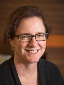 A woman with glasses and curly brown hair smiles warmly against a blurred brown background.