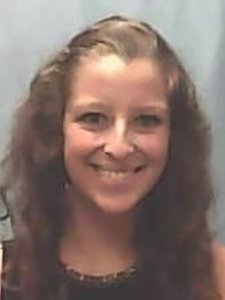A woman with long wavy brown hair smiles at the camera against a plain light blue background.