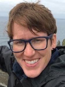 A person with short brown hair and dark glasses smiles at the camera outdoors near a cliff and ocean.