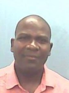 A man with short hair wearing a light pink collared shirt is looking at the camera against a plain light blue background.