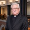 A man with white hair and glasses wearing a black clerical collar and suit jacket stands indoors near a railing.