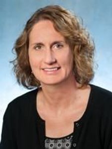 A woman with shoulder-length curly light brown hair wearing a black cardigan over a patterned top smiles against a blue.