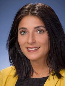A woman with shoulder-length dark hair smiles slightly against a blue background wearing a yellow jacket and black top.