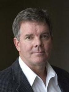 A middle-aged man with short gray hair wearing a white shirt and dark blazer looks directly at the camera against a dark.