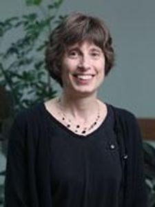 A woman with short brown hair wearing a black top and necklace smiles in front of a blurred green plant.