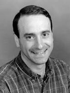 A man with short dark hair smiles at the camera wearing a plaid shirt against a plain background.