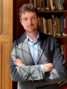 A JCU faculty member with curly brown hair and a beard, wearing a blazer, poses with crossed arms in front of a bookshelf.