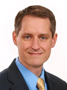 A man with short brown hair wearing a blue shirt, yellow patterned tie, and dark suit jacket smiles slightly against a white.