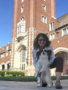 A woman with curly dark hair and glasses smiles while standing indoors near a railing with blurred background.