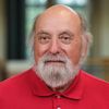 An older man with a bald head and white beard wearing a red polo shirt looks directly at the camera.