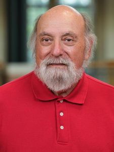 An older man with a bald head and white beard wearing a red polo shirt looks directly at the camera.