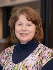A woman with light brown hair wearing a colorful patterned top over a dark turtleneck smiles softly at the camera.