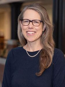A woman with long gray hair, glasses, and a pearl necklace smiles while wearing a dark top in an indoor setting.