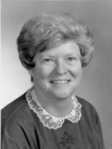 A black and white portrait of a smiling woman with short curly hair wearing a dark top with a white lace collar.