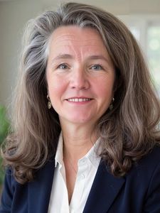 A woman with shoulder-length gray hair wearing a white blouse and dark blazer smiles in a bright room with plants.