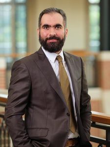 A man with a beard and mustache wearing a brown suit and gold tie stands indoors near a railing, looking at the camera.