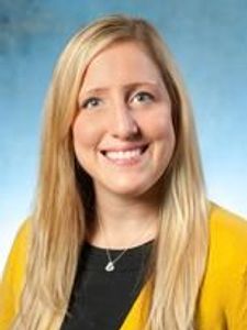A smiling woman with long blonde hair wearing a black top and yellow cardigan against a blue background.