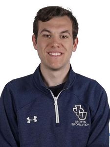 A young man with short dark hair smiles wearing a navy blue quarter-zip jacket with a sports logo on a white background.