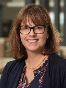 A woman with shoulder-length brown hair and glasses smiles while wearing a navy cardigan and patterned blouse indoors.