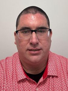 A man with short dark hair and glasses wearing a red patterned shirt is looking directly at the camera against a plain light.