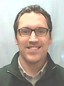 A man with short dark hair and glasses smiles while wearing a dark jacket over a collared shirt against a plain background.