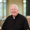 An older man with white hair wearing a black clerical shirt with a white collar is smiling indoors.