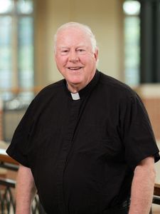 An older man with white hair wearing a black clerical shirt with a white collar is smiling indoors.