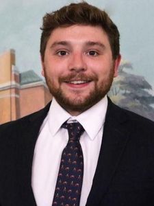 A man with short brown hair and a beard smiles wearing a dark suit, white shirt, and patterned tie against a painted.