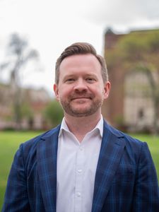 A man with short brown hair and a beard wearing a blue plaid jacket and white shirt stands outdoors on green grass.