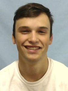 A young man with short brown hair smiles slightly while wearing a white shirt against a plain light blue background.
