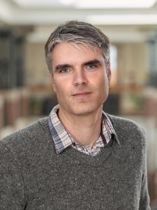 A man with gray hair and blue eyes wears a gray sweater over a plaid shirt and looks directly at the camera.