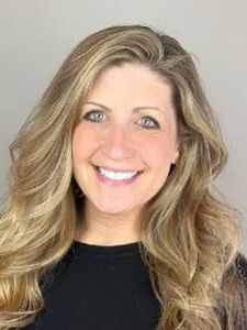 A woman with long wavy blonde hair smiles warmly against a plain light gray background.